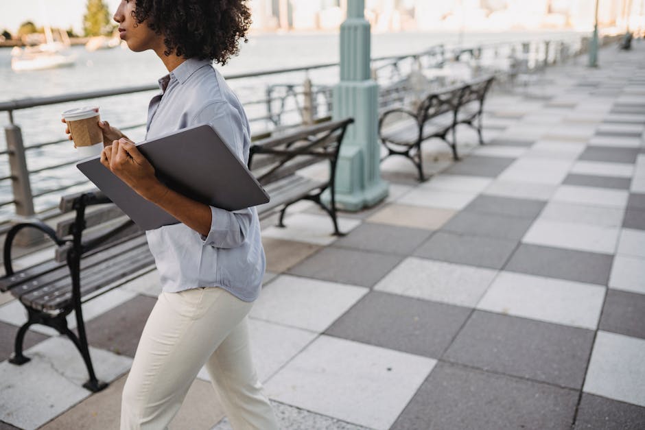 A woman strolls along a riverside promenade, holding a laptop and coffee cup.