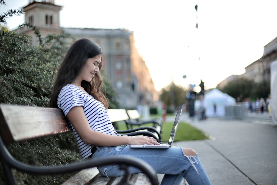 Woman sitting on a bench in a city park, working remotely on a laptop during the day.