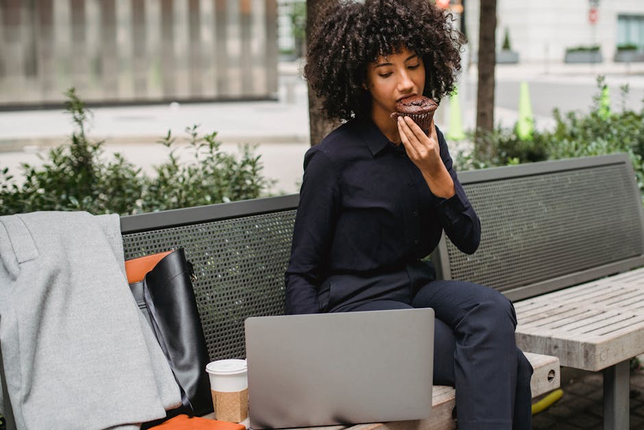 Businesswoman enjoys a muffin on an outdoor bench with a laptop and coffee.