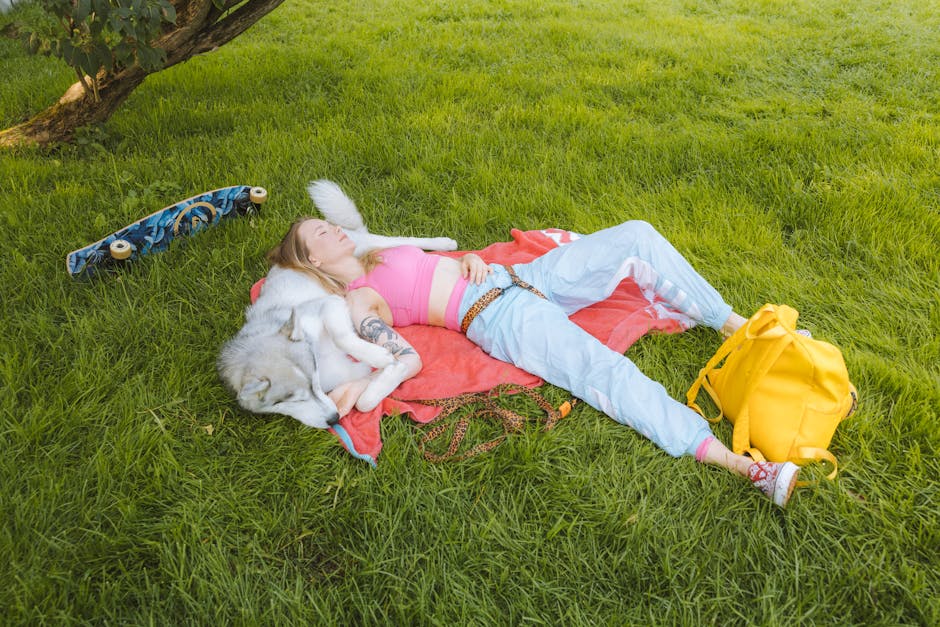 A young woman in a pink top relaxes with her husky dog on a picnic blanket in a sunny park.